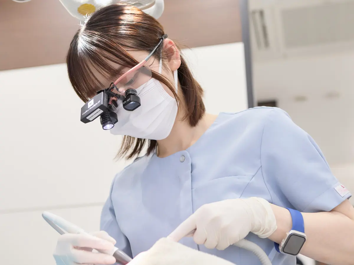 ライト付きルーペを装着した歯科衛生士が、器具を手に患者さんの処置を行っている様子。水色のスクラブに白い手袋
