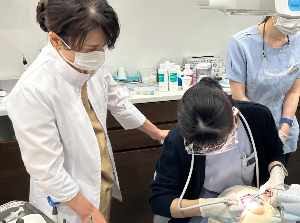 一流の外部講師がユニットサイドでDHにマンツーマンの実技指導をしている場面。スタッフが真剣な表情で手元を見ている。いつもの診療室での指導というリアルさが重要。