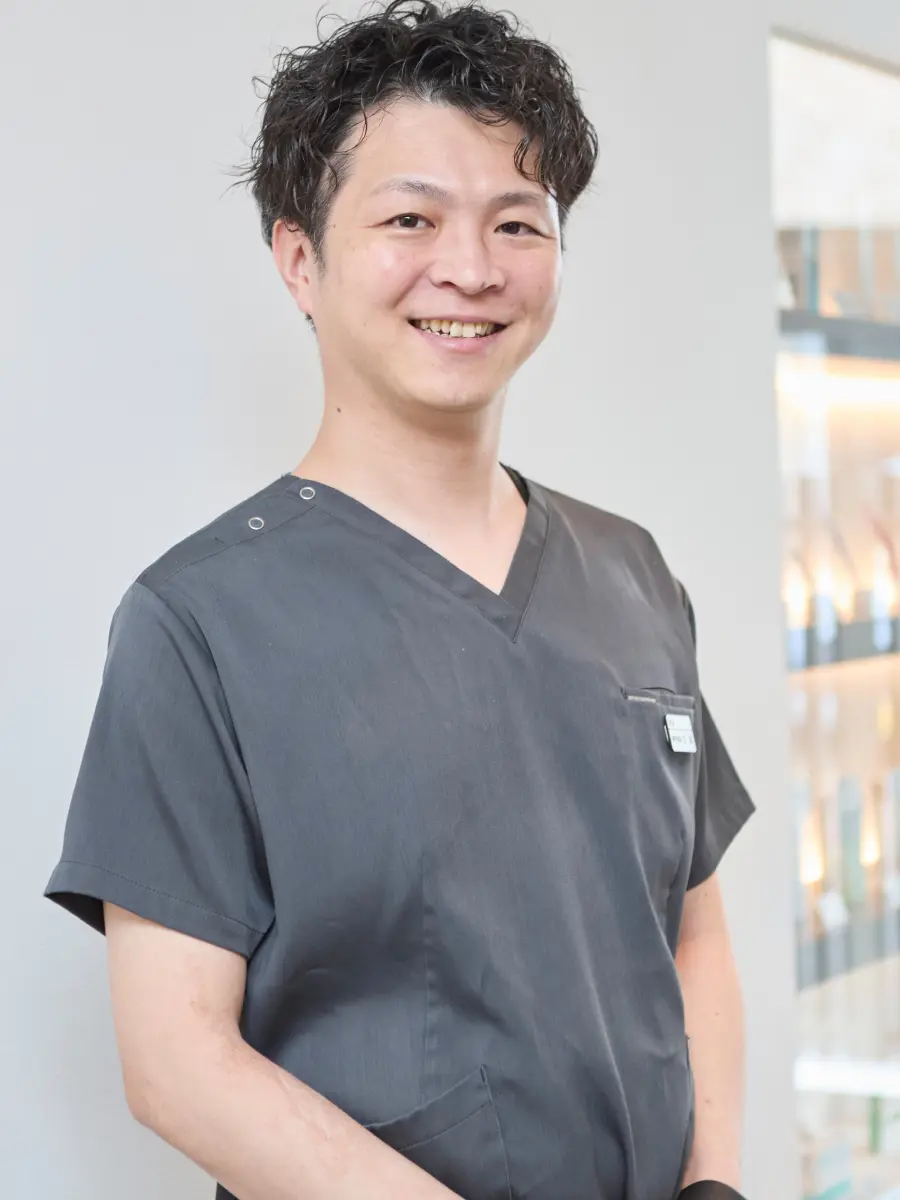 グレーのスクラブを着た院長が、正面を向いて笑顔で立っている縦位置のポートレート