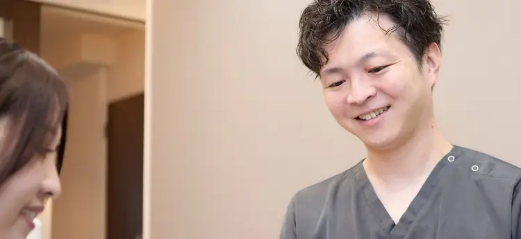 グレーのスクラブを着た院長が、笑顔で目の前の相手に語りかけている表情のアップ。左手前にスタッフの後ろ姿